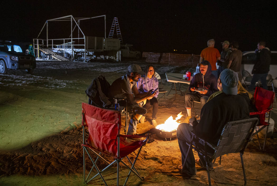 Friends hang out and warm up by a gas fire pit near the main stage still under construction as ...