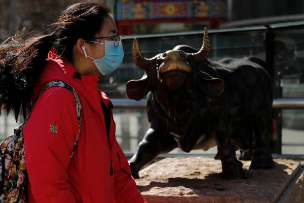 A woman wearing a protective face mask walks by the investment icon bull statue on display outs ...