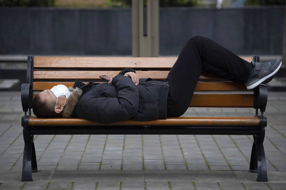 A man wearing a mask to help protect against the spread against the coronavirus naps on a bench ...