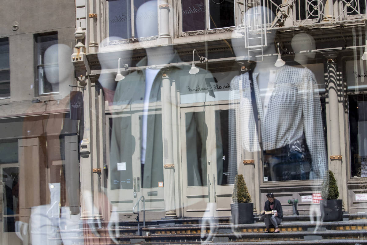 A woman sitting on a stoop reading a book in the sun is seen reflected by a closed clothing sto ...