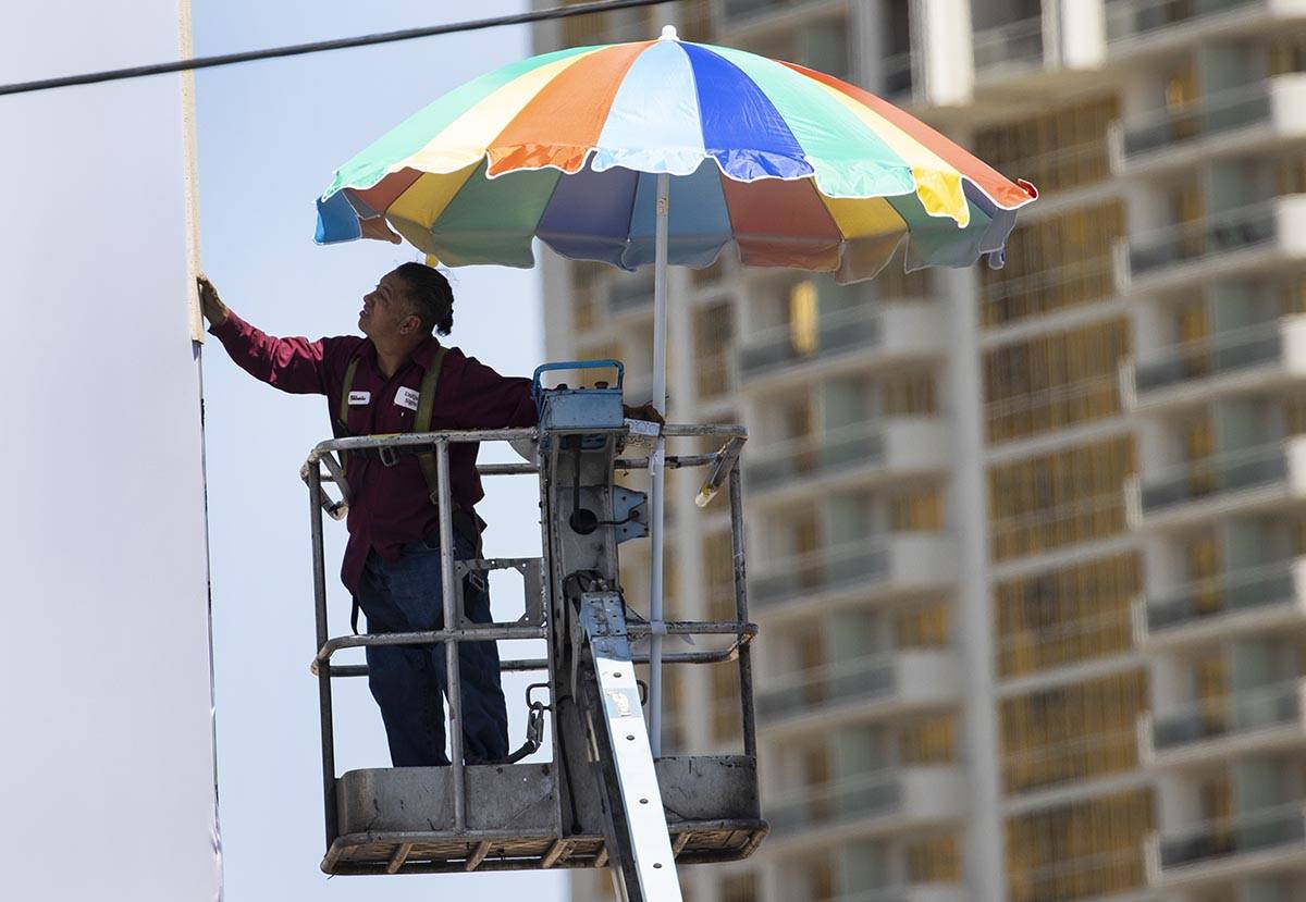 A worker uses a giant umbrella to protect himself from sun as he works on an outdoor advertisin ...