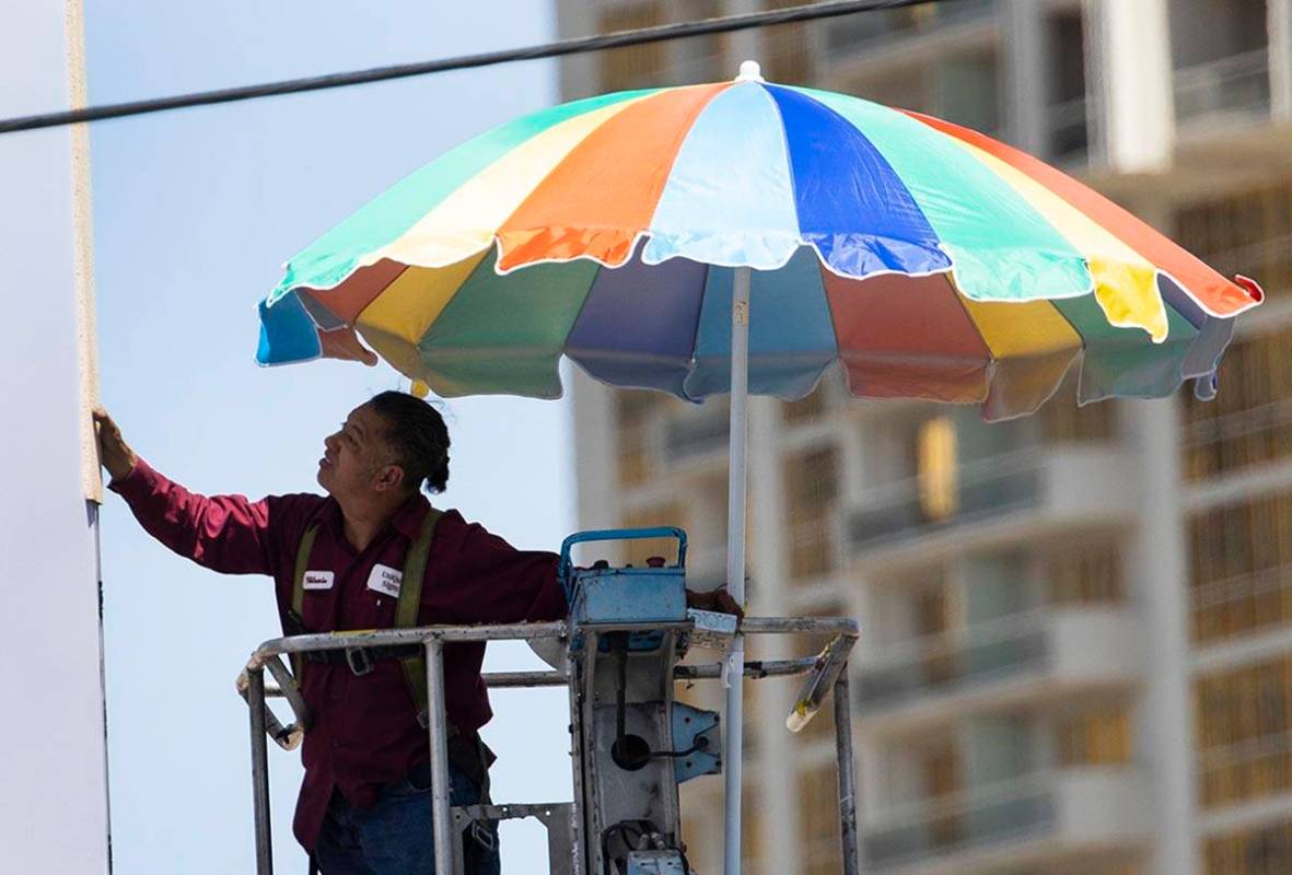 A worker uses a giant umbrella to protect himself from sun as he works on an outdoor advertisin ...