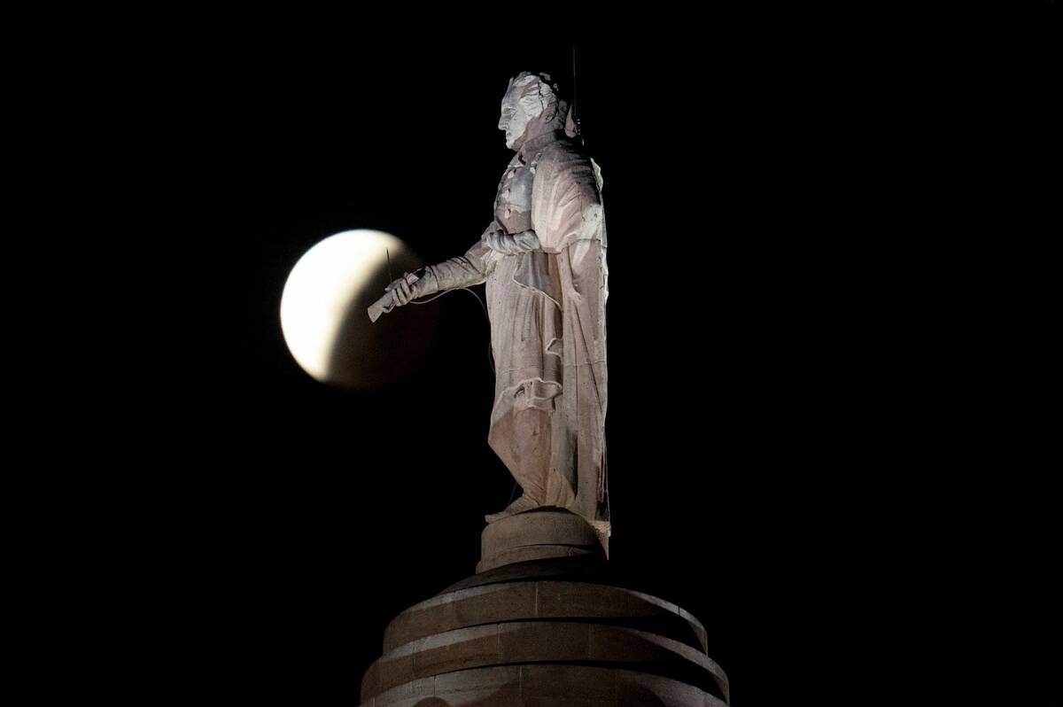 The earth's shadow covers the full moon during a partial lunar eclipse visible near a statue of ...