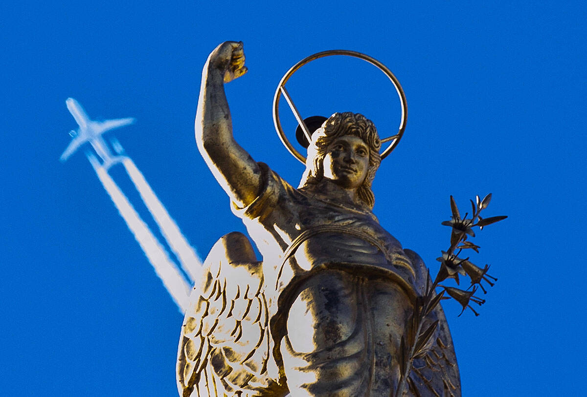 A passenger plane flies in clear blue sky and leaves a white trail as it flies behind a statue ...
