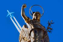 A passenger plane flies in clear blue sky and leaves a white trail as it flies over a statue at ...