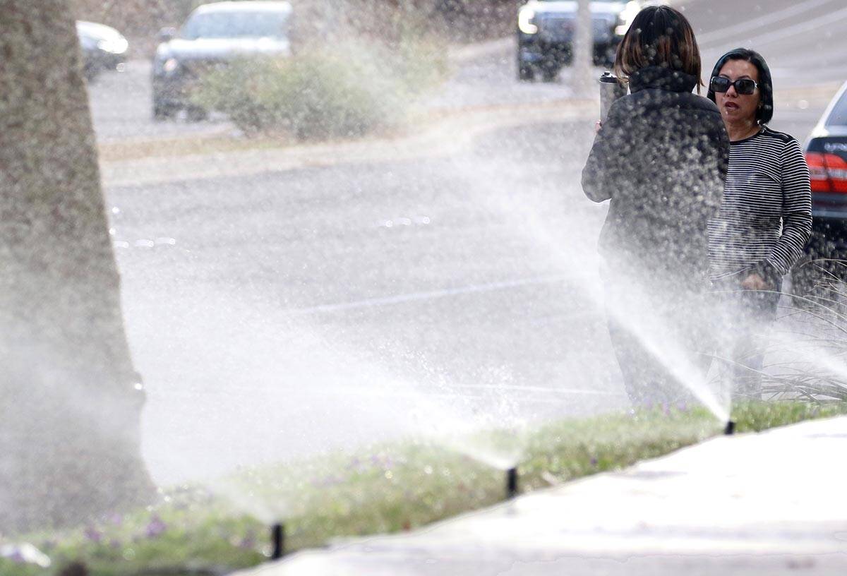 Pedestrians wait to cross Green Valley Parkway as lawn sprinklers water grass in March 2019 in ...