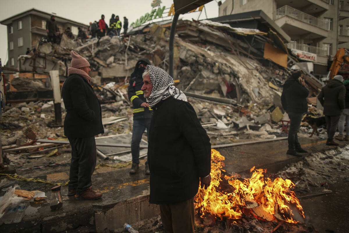 People warm themselves next to a collapsed building in Malatya, Turkey, Tuesday, Feb. 7, 2023. ...