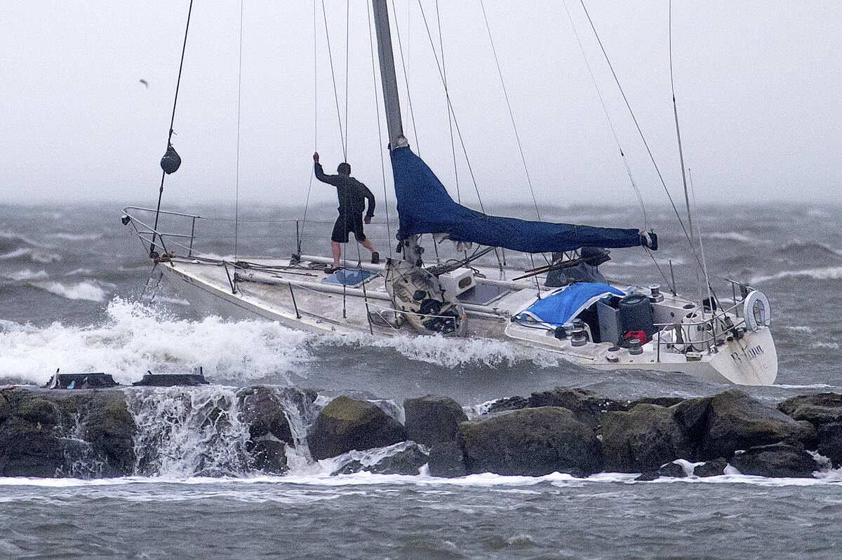 A boater stands on the bow of an anchored sailboat as waves crash over a breakwater in Alameda, ...