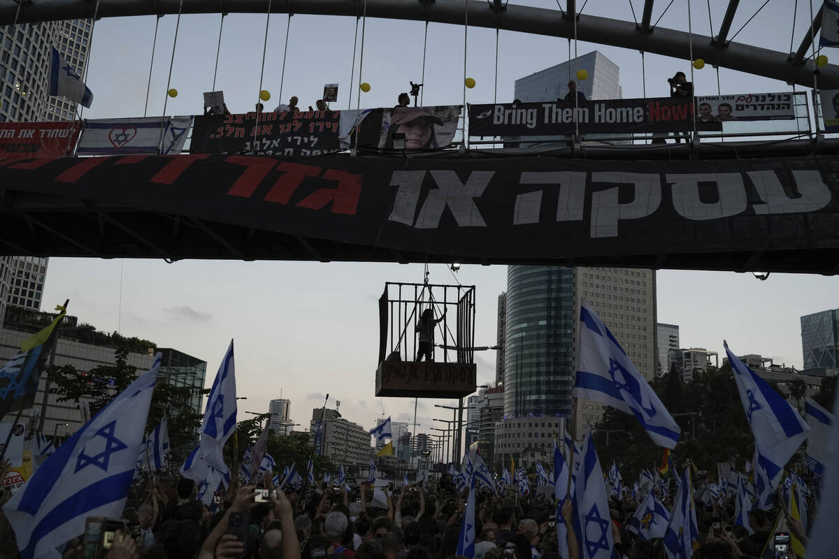 A demonstrator stands in a cage hanging from a pedestrian bridge as others wave Israeli flags d ...