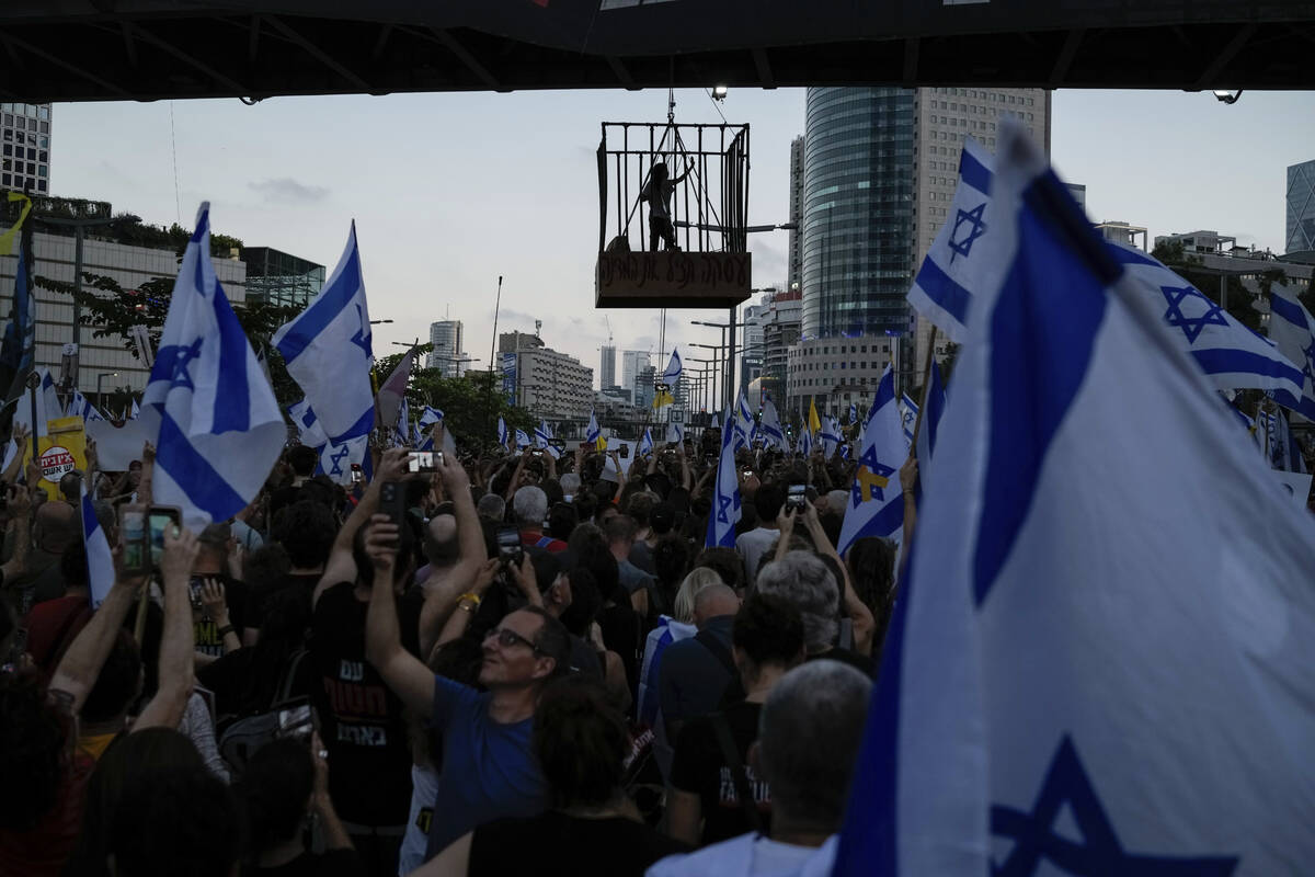 A demonstrator stands in a cage hanging from a pedestrian bridge as others wave Israeli flags d ...