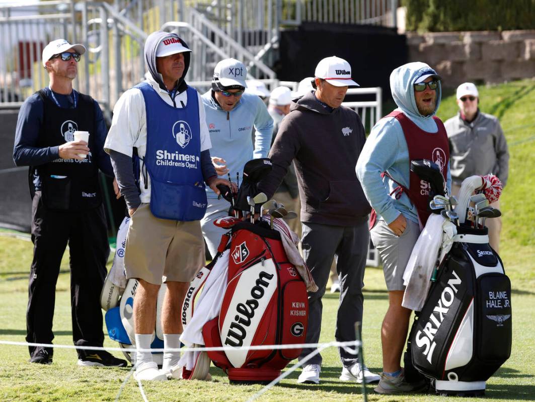 Players and their caddies are bundled up to protect themselves from heavy wind as they wait to ...