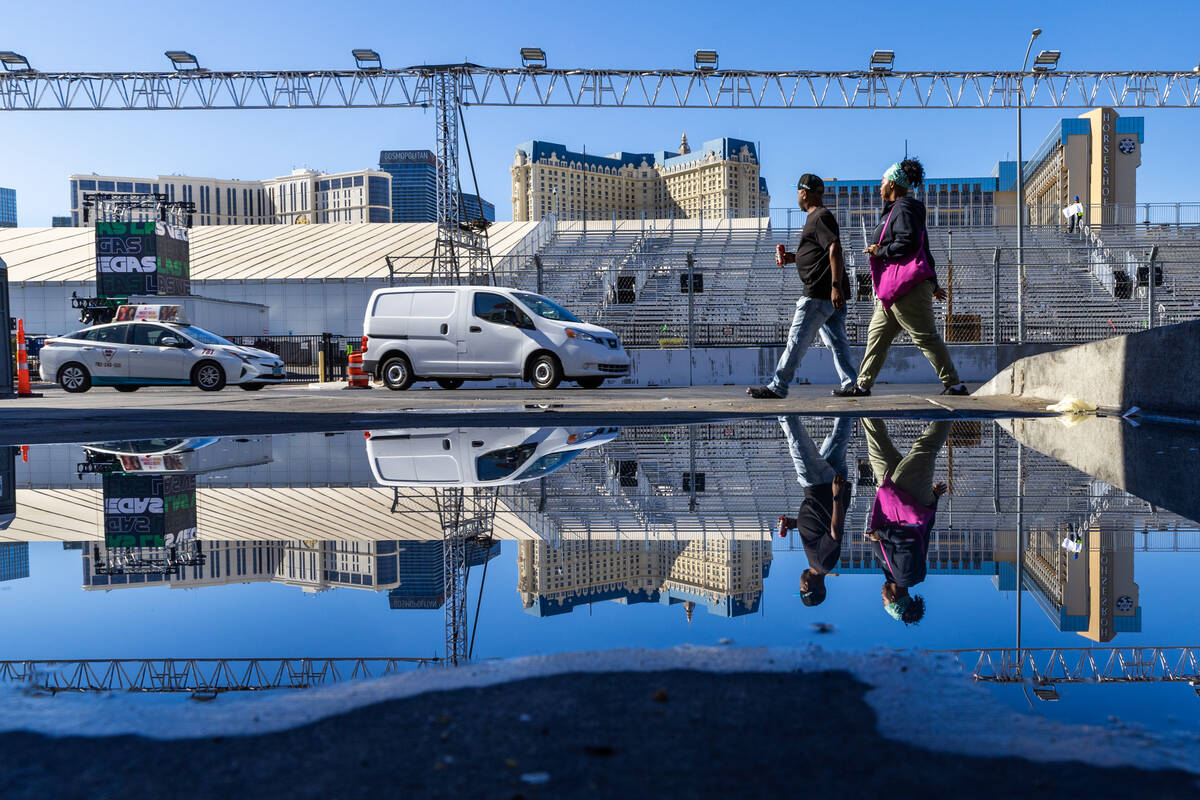 Pedestrians are reflected in a large puddle as grandstands and race barriers are now in along K ...